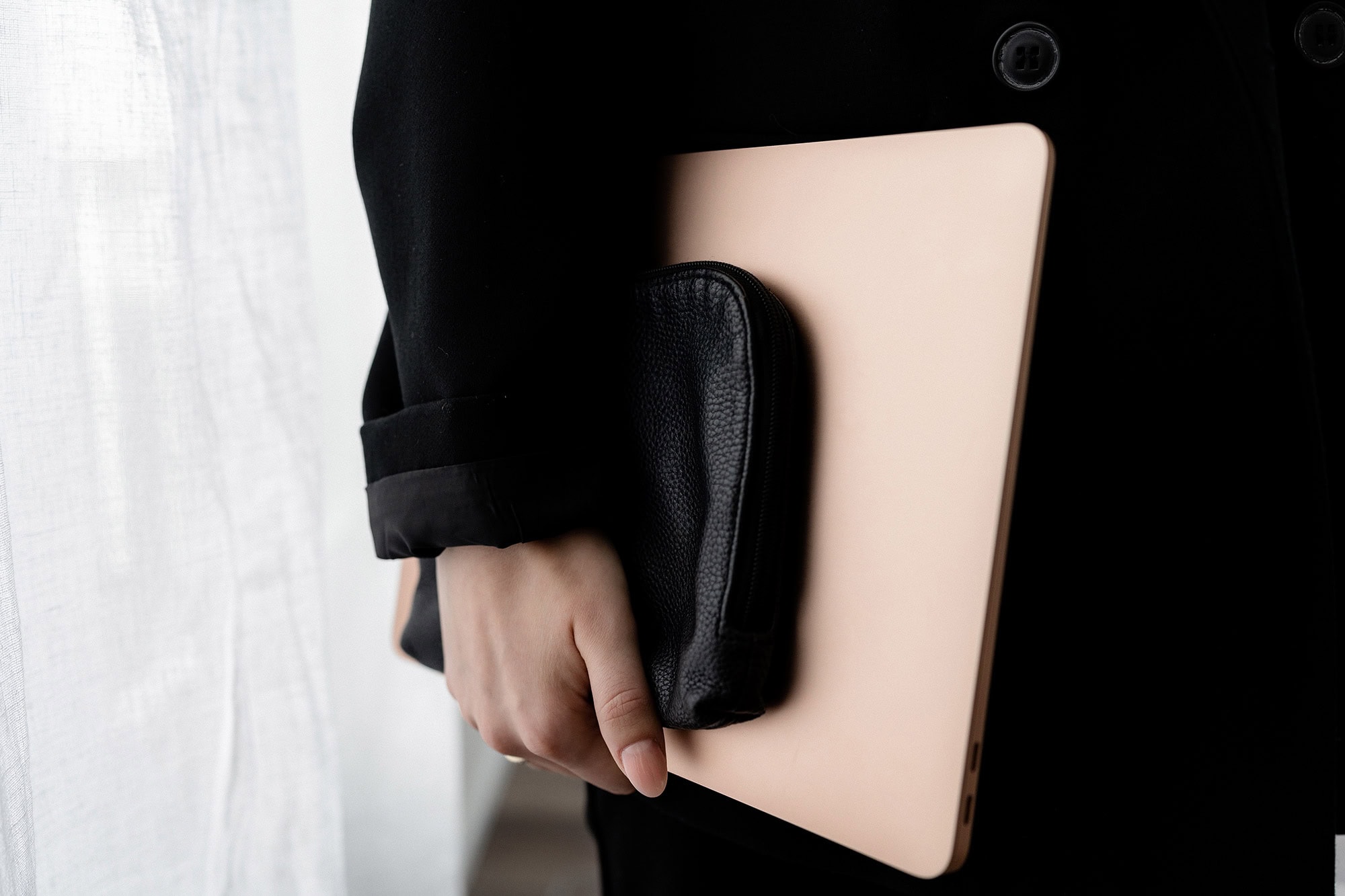 Eine Person in einem schwarzen Mantel hält einen goldfarbenen Laptop und eine kleine schwarze Ledertasche an ihrer Seite und steht neben einem weißen Vorhang.