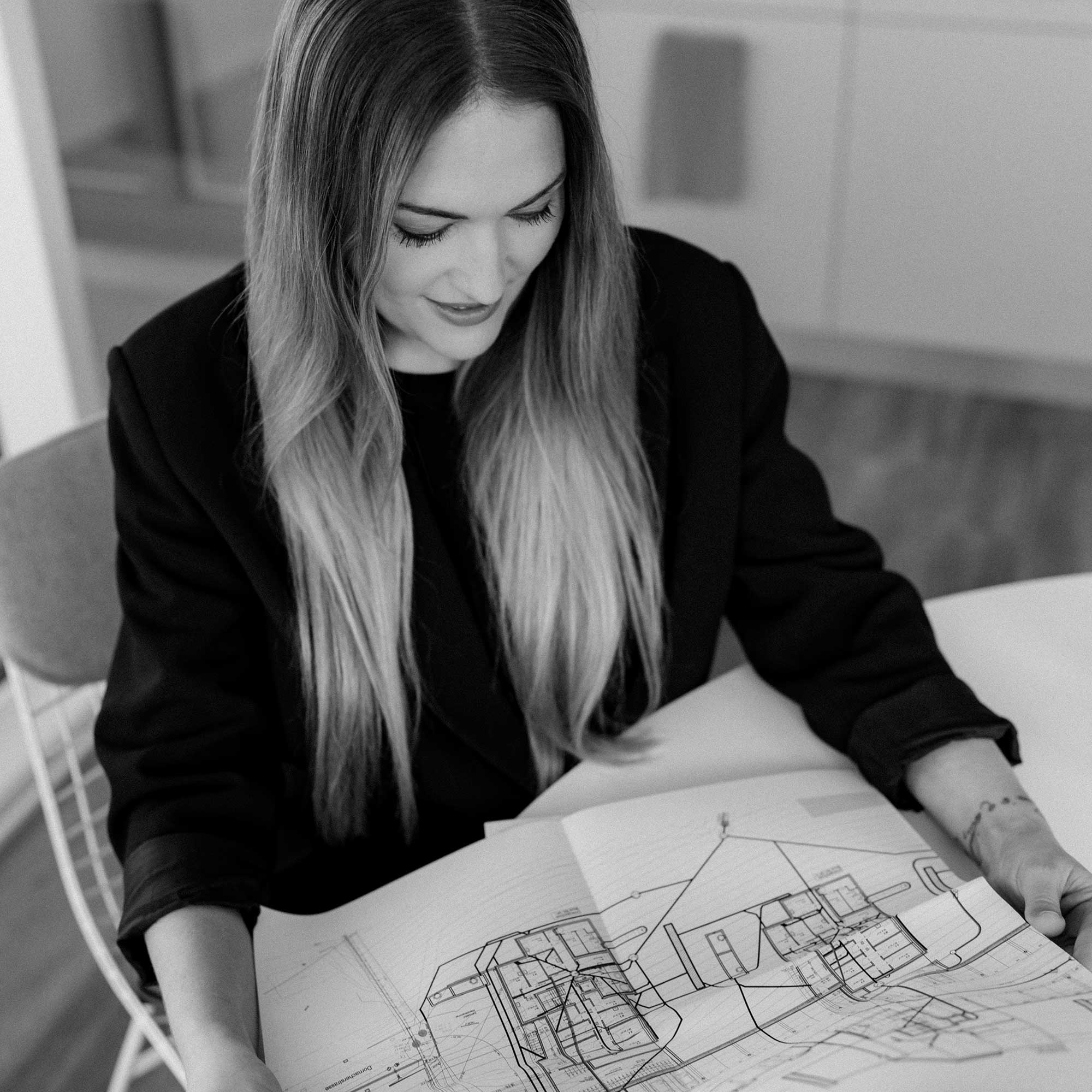 Eine Frau mit langem, glattem Haar sitzt an einem Tisch und blickt lächelnd auf die vor ihr ausgebreiteten Baupläne. Das Bild ist in schwarz-weiß gehalten.