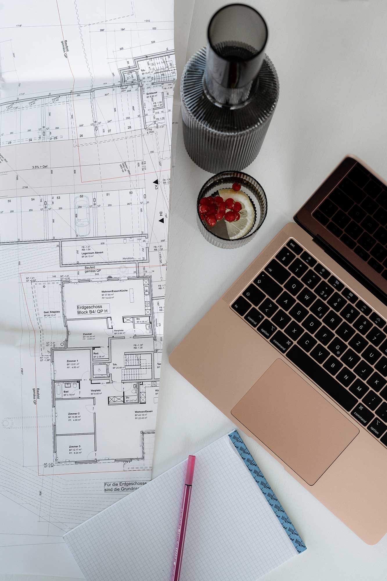 Ein ausgerollter architektonischer Grundriss liegt neben einem roségoldenen Laptop, einem Notizblock mit rosa Stift und Granatapfelkernen auf einem weissen Schreibtisch - und fängt den kreativen Prozess der Erstvermietung an der Hofackerstrasse 10 in Muttenz ein.