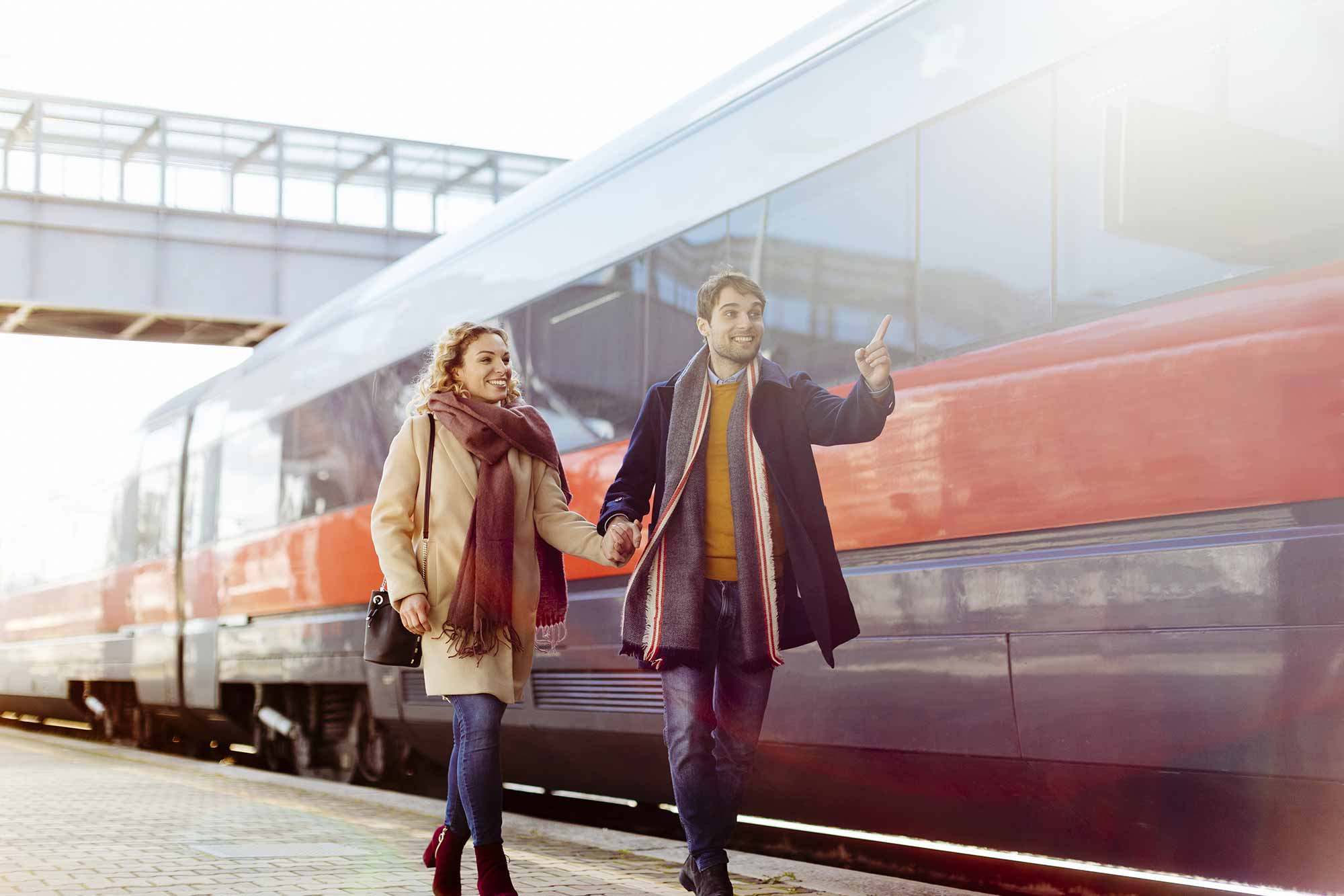 Ein lächelndes Paar in Winterkleidung geht Hand in Hand über einen Bahnsteig in der Nähe der Hofackerstrasse 10 in Muttenz, mit einem modernen Zug hinter ihnen. Der Mann gestikuliert aufgeregt, während die Frau nach vorne schaut. Beide wirken glücklich und entspannt. Ideal für die Erstvermietung.