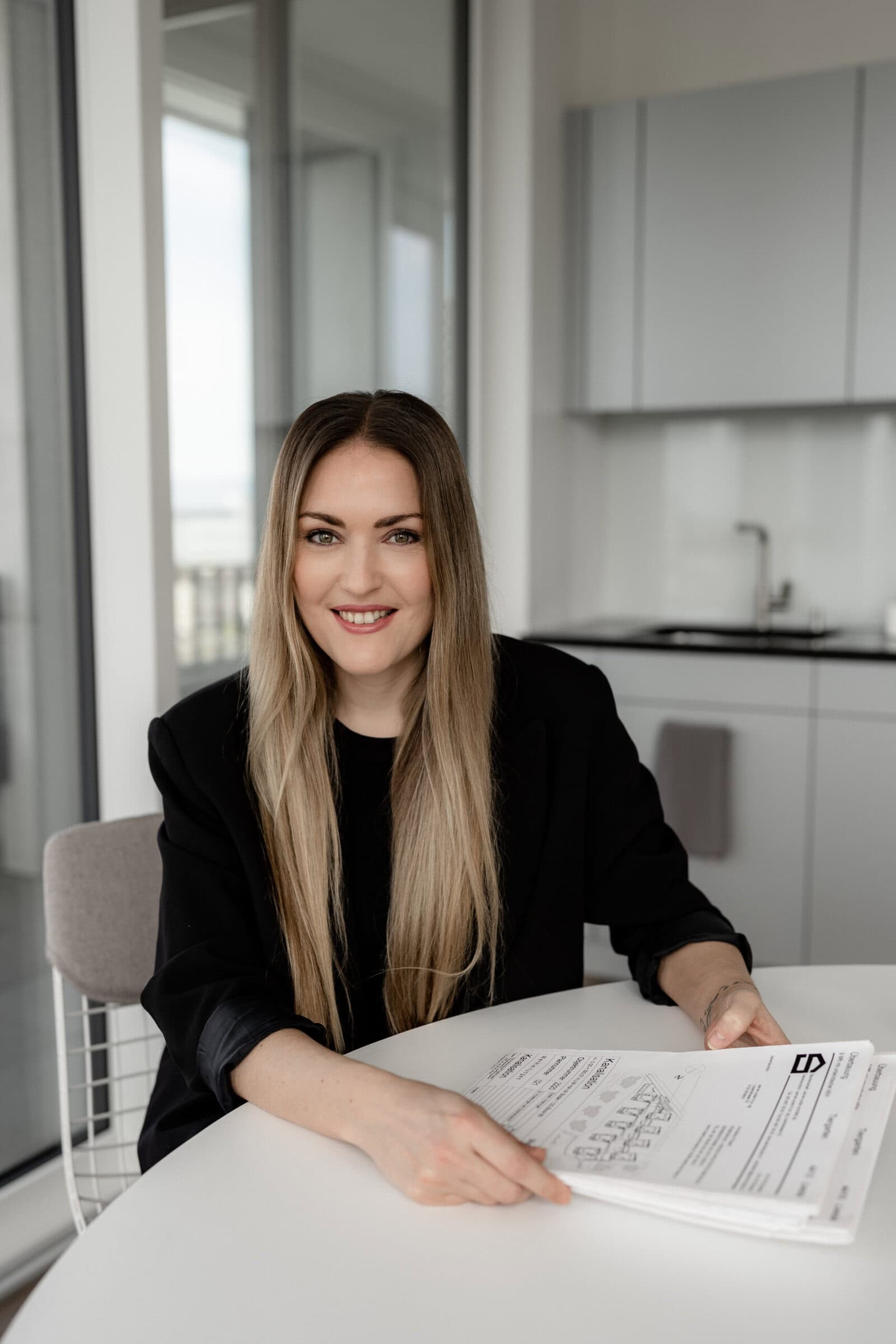 Eine Frau mit langen blonden Haaren, die einen schwarzen Blazer trägt, sitzt an einem Tisch, hält Papiere in der Hand und lächelt. Sie befindet sich in einem modernen, lichtdurchfluteten Büro von Kokon Immobilien mit Schränken und großen Fenstern im Hintergrund.